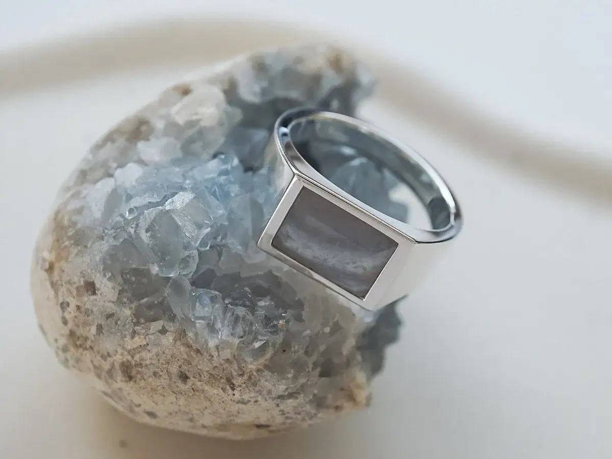 Ein silberner Ring mit einem rechteckigen, grau-weißen Schmuckstein ruht auf einem blauen Kristall.
