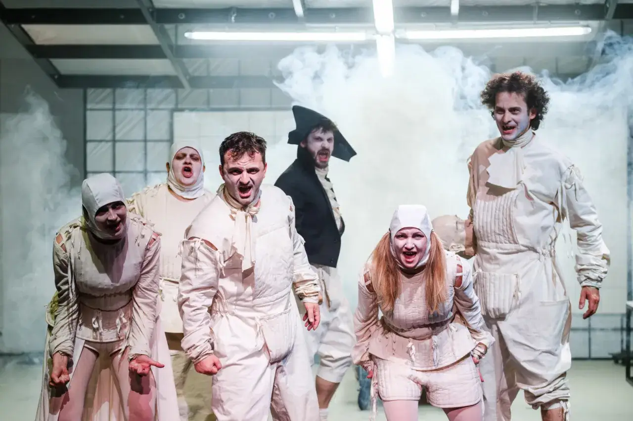 Aktorzy w białych strojach, z przerażonymi minami, tworzą scenę pełną grozy i komizmu w obrazie śmierci. Dym i teatralne światła podkreślają nastrój.