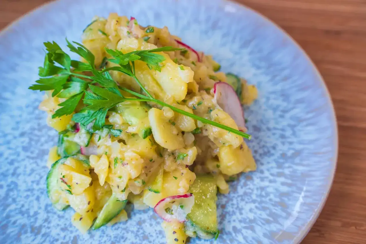 Ein köstlicher Kartoffelsalat mit Gurken, Radieschen und frischer Petersilie auf einem blauen Teller.