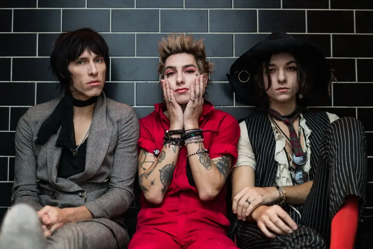 Trzech członków Palaye Royale w stylowych, rockowych strojach siedzi obok siebie na tle czarnych płytek.