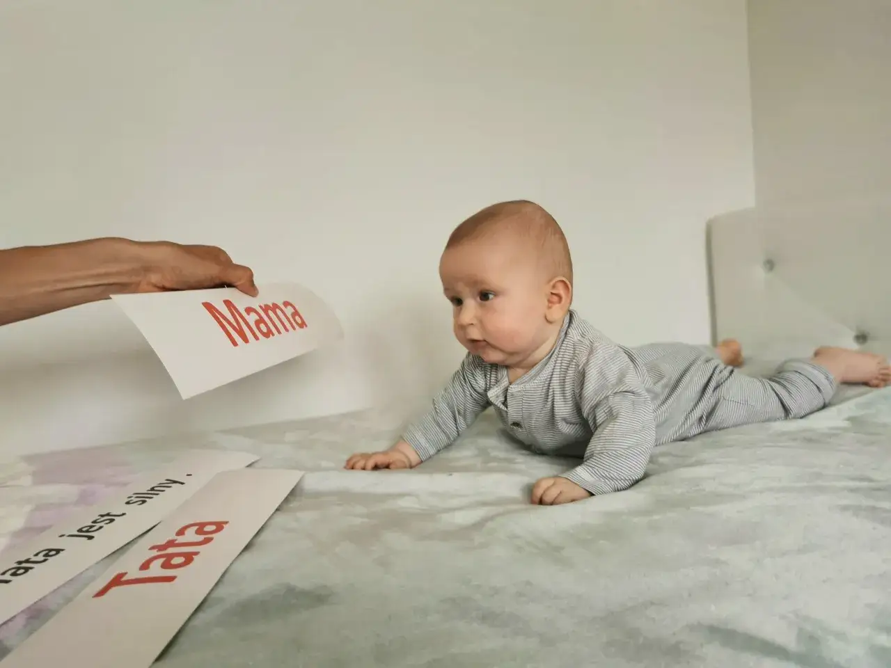 Zaciekawione niemowlę na brzuchu patrzy na kartkę z napisem "Mama". Obok leżą kartki "Tata" i "Tata jest silny".