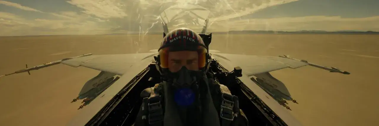 Pilot myśliwca w hełmie z napisem "Maverick" w kokpicie, lecący nad pustynnym krajobrazem.