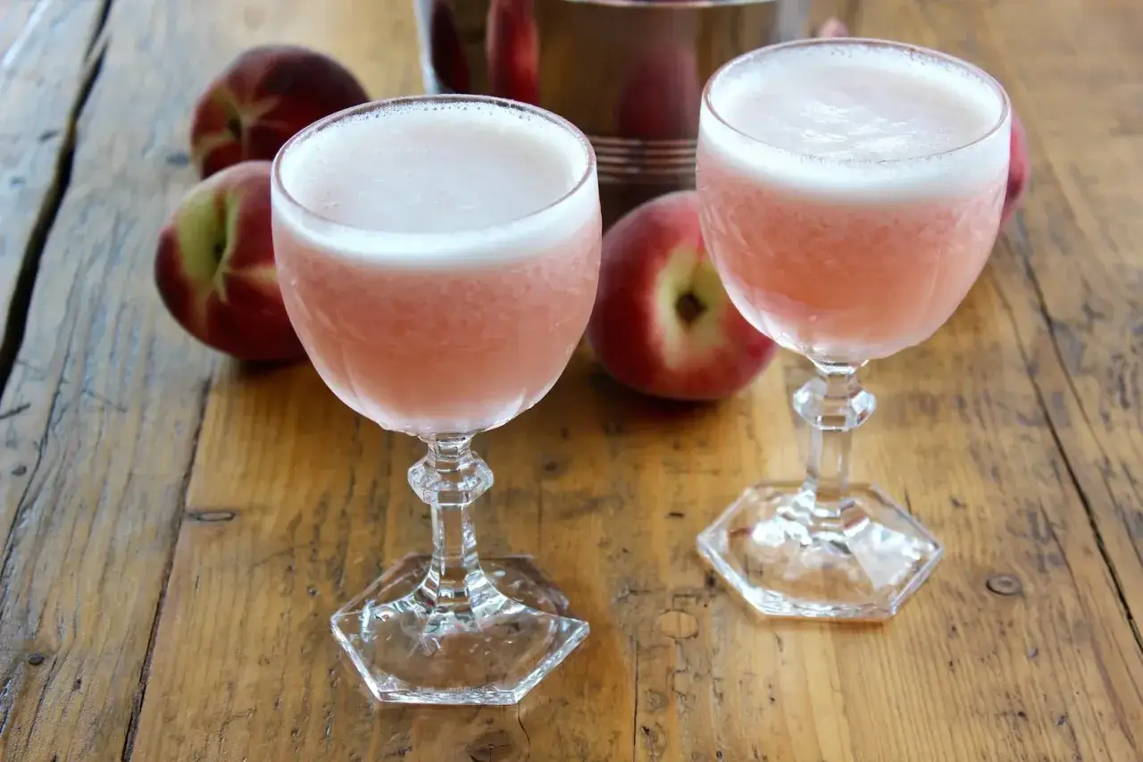 Dwa różowe drinki z pianką, obok brzoskwiń, na drewnianym stole. To jest właśnie co to znaczy drink – orzeźwienie i smak.