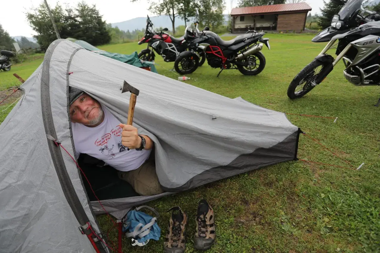 Mężczyzna w namiocie z siekierką, obok motocykle. Warto zabrać na wyprawę motocyklową namiot, buty i siekierkę.