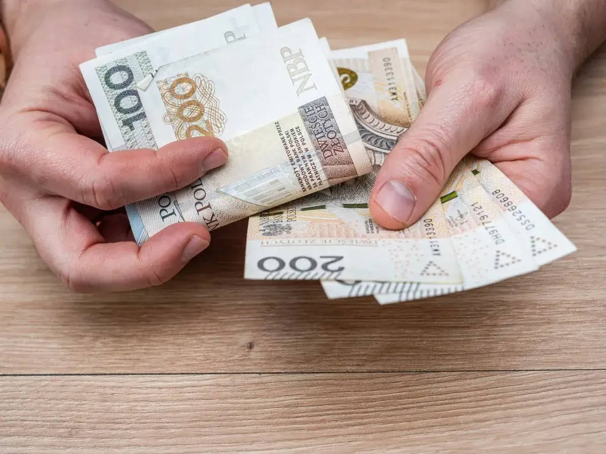 Dłonie trzymają polskie banknoty 100 i 200 zł. Może to być pomoc w opłaceniu rachunków, np. bon ciepłowniczy.