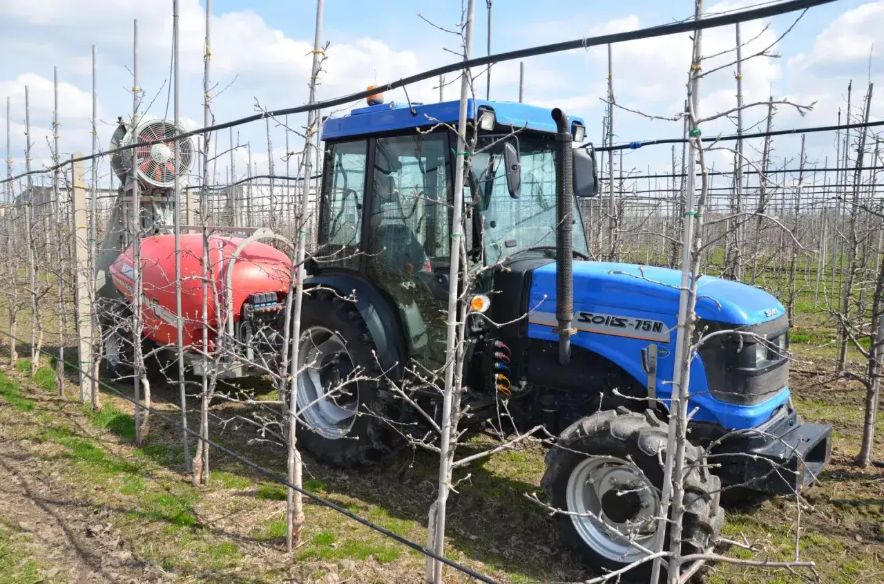 Niebieski traktor z opryskiwaczem w sadzie, przygotowany do zabiegu środkiem ochrony roślin, np. acetamiprydem, chroniącym młode drzewka.
