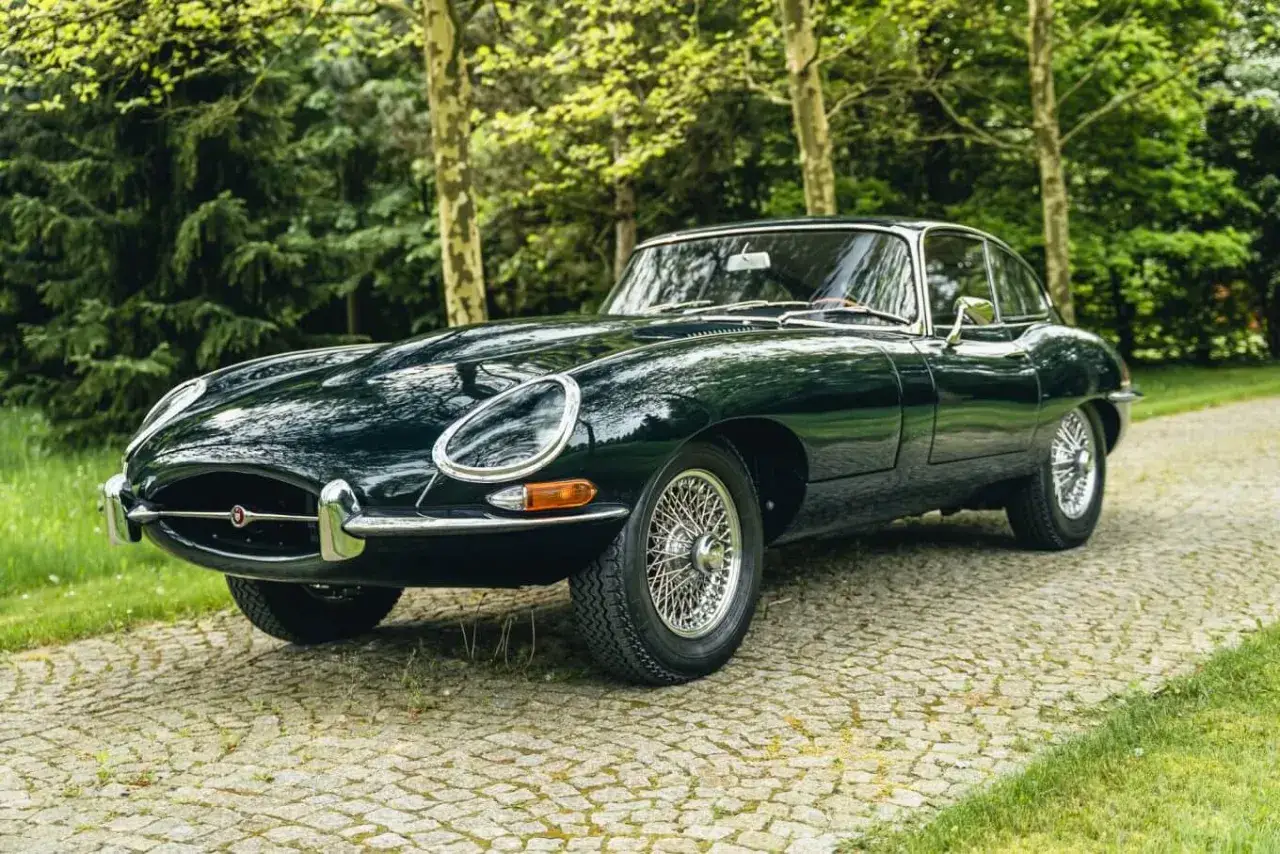 Klasyczny, ciemnozielony Jaguar E-Type na brukowanej drodze, otoczony drzewami.