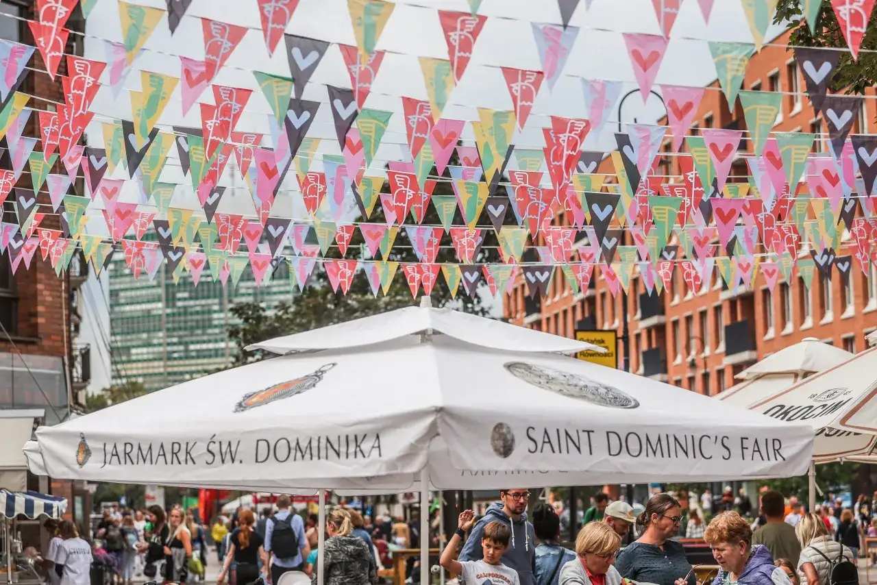 Kolorowe chorągiewki ozdabiają ulicę podczas Jarmarku Świętego Dominika. Ludzie spacerują pod białymi parasolami z napisami "Jarmark Św. Dominika" i "Saint Dominic's Fair".