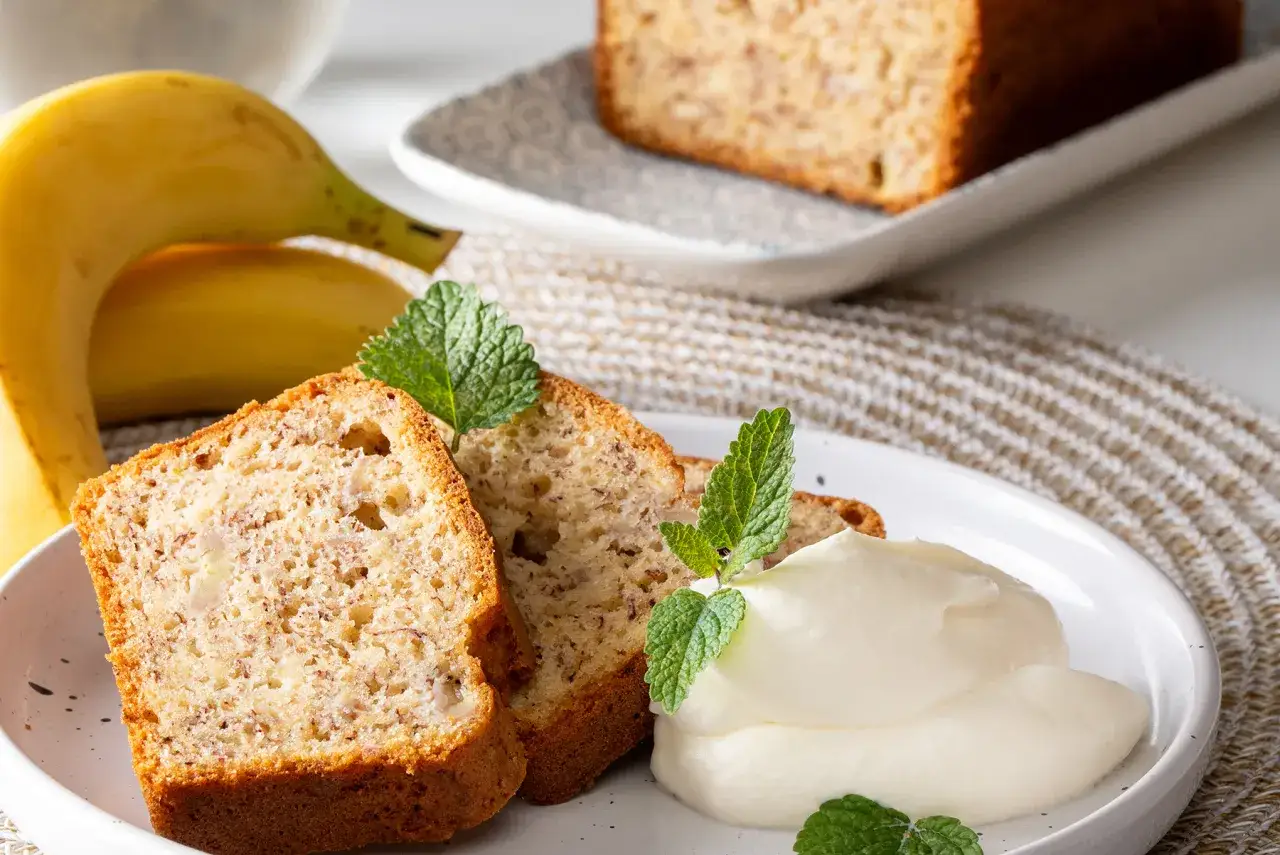 Slices of banana bread with cream and mint leaves, next to whole bananas.