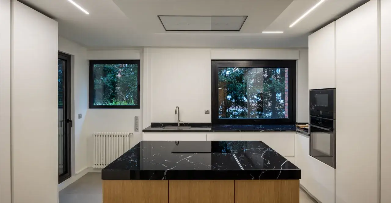 Cocina moderna con **fregadero en isla** de mármol negro y encimeras a juego. Muebles blancos y electrodomésticos integrados.