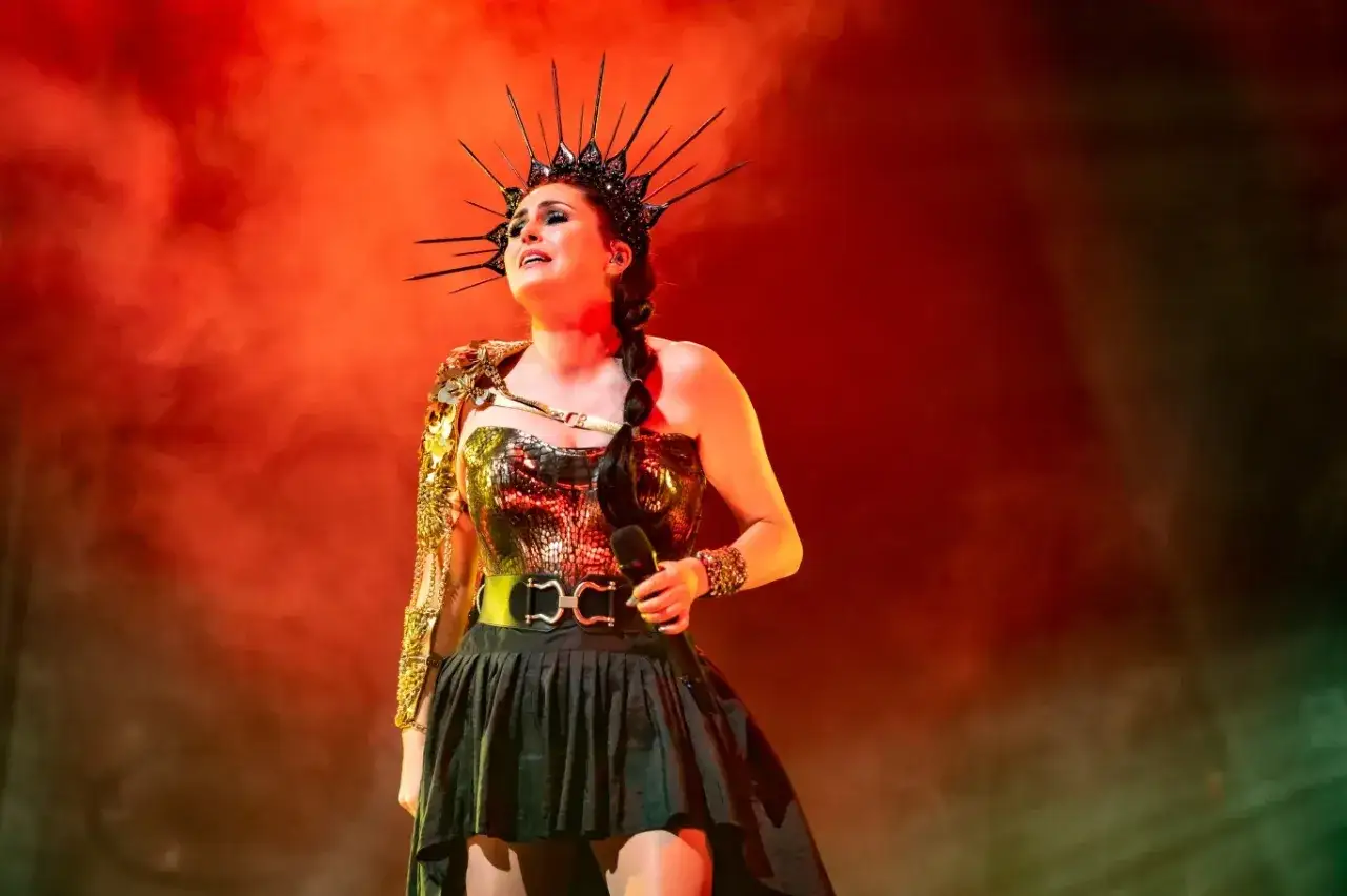 Wokalistka Within Temptation w koronie i błyszczącej kreacji, z mikrofonem w dłoni, na tle czerwonej mgły.