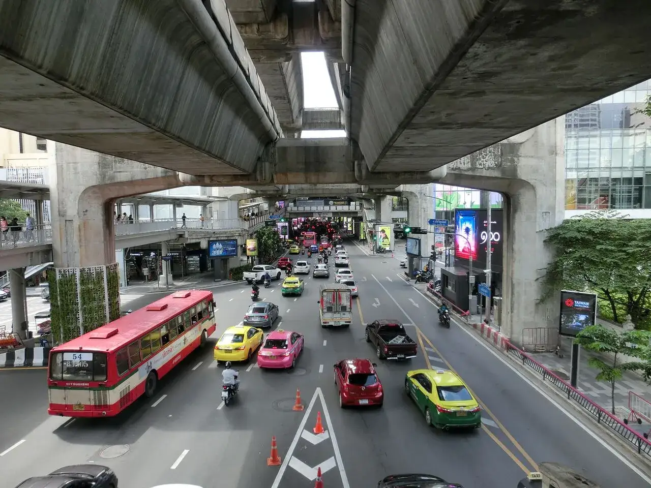 Tętniący życiem transport w Bangkoku: czerwony autobus, żółte i różowe taksówki, samochody i motocykle na ruchliwej ulicy pod betonową estakadą.