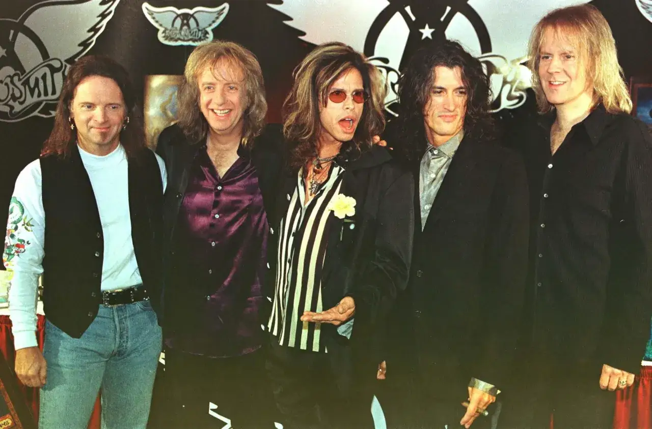 Steven Tyler mlody z zespołem Aerosmith, w charakterystycznych okularach i stroju, gotowy na scenę.