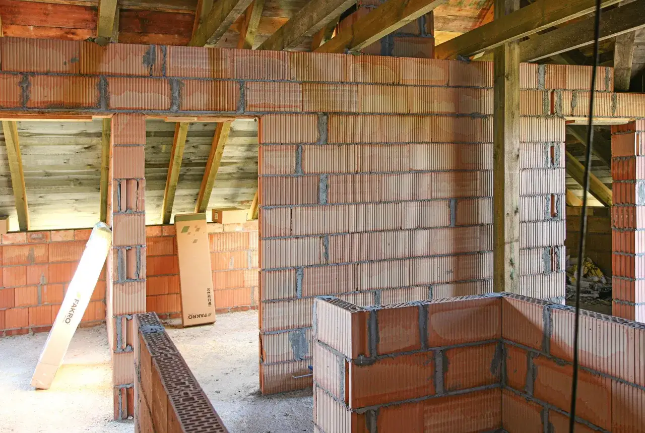 Budowa domu z bloczków z betonu komórkowego. Widać ściany z pustaków, drewniane belki stropowe i materiały budowlane, jak klej czy zaprawa do betonu komórkowego.