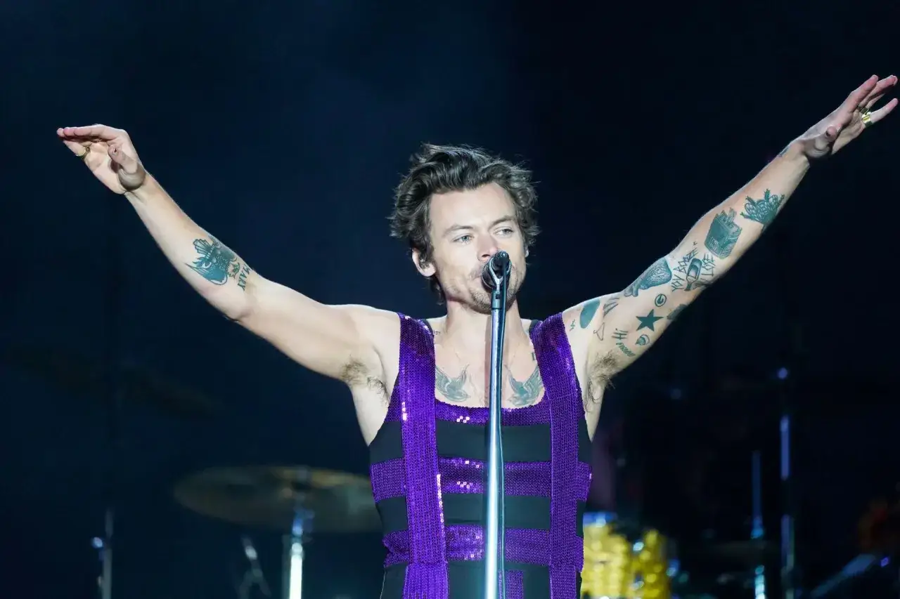 Harry Styles w błyszczącym fioletowym topie śpiewa na koncercie, unosząc ramiona. Jego ramiona pokryte są tatuażami.