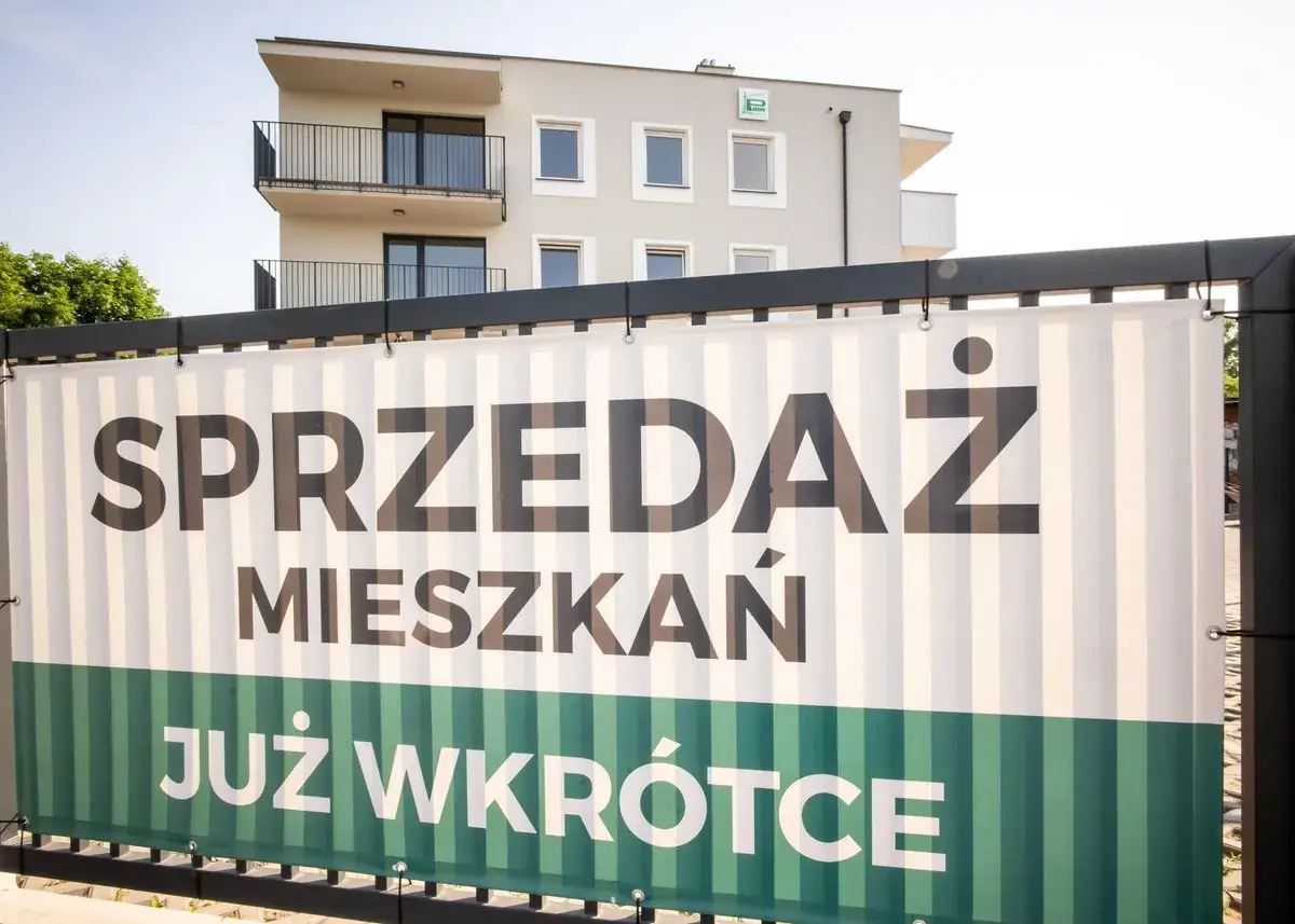 Baner z napisem "SPRZEDAŻ MIESZKAŃ JUŻ WKRÓTCE" na tle nowoczesnego budynku mieszkalnego.