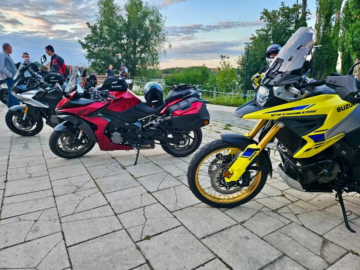 Kilka motocykli Suzuki zaparkowanych na brukowanym placu, z ludźmi w tle.