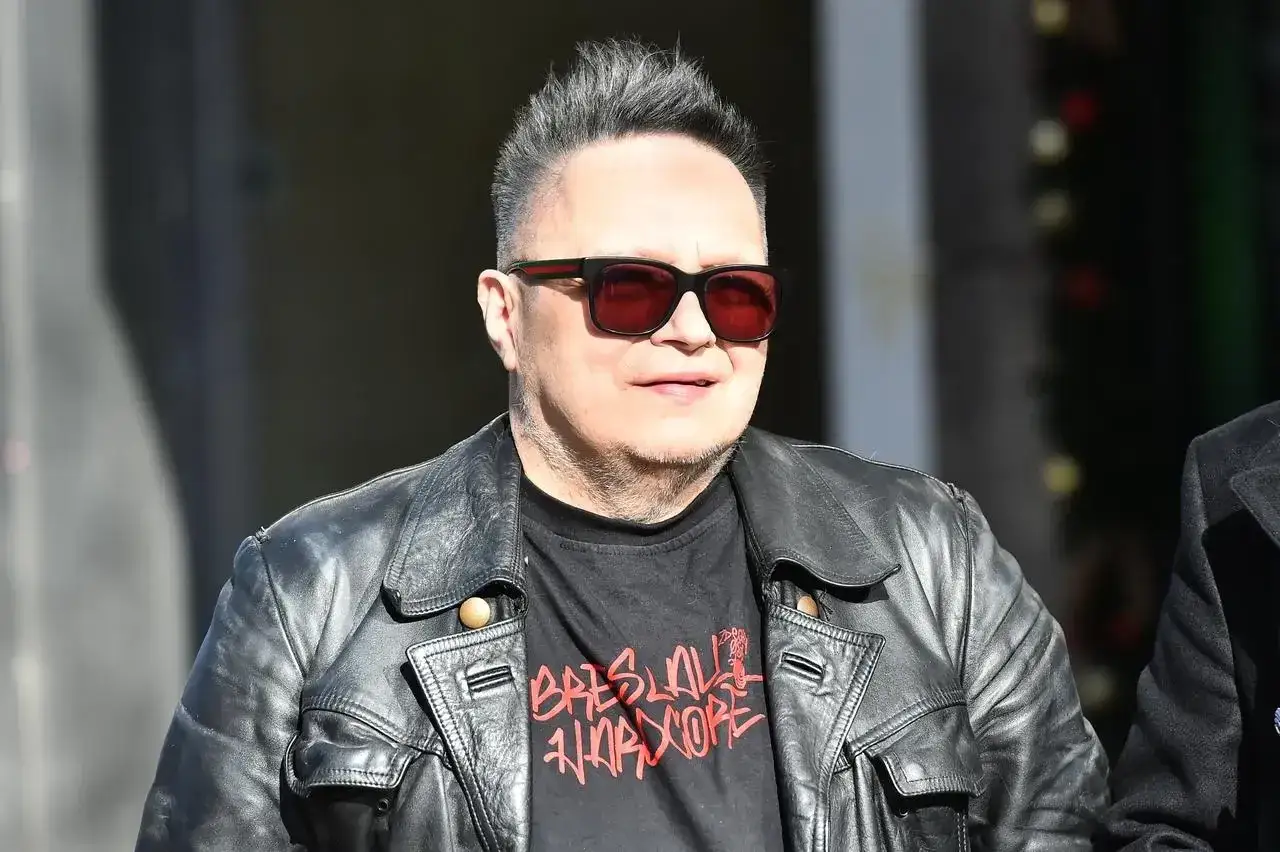 Charyzmatyczny wokalista w skórzanej kurtce i okularach przeciwsłonecznych, z charakterystyczną fryzurą.