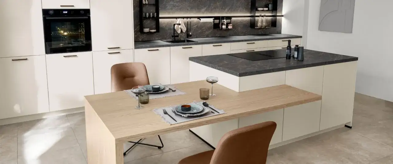 Cocina moderna con encimeras de piedra oscura y gabinetes blancos. La mesa de madera clara y las sillas de cuero marrón complementan los materiales para cocinas modernas.
