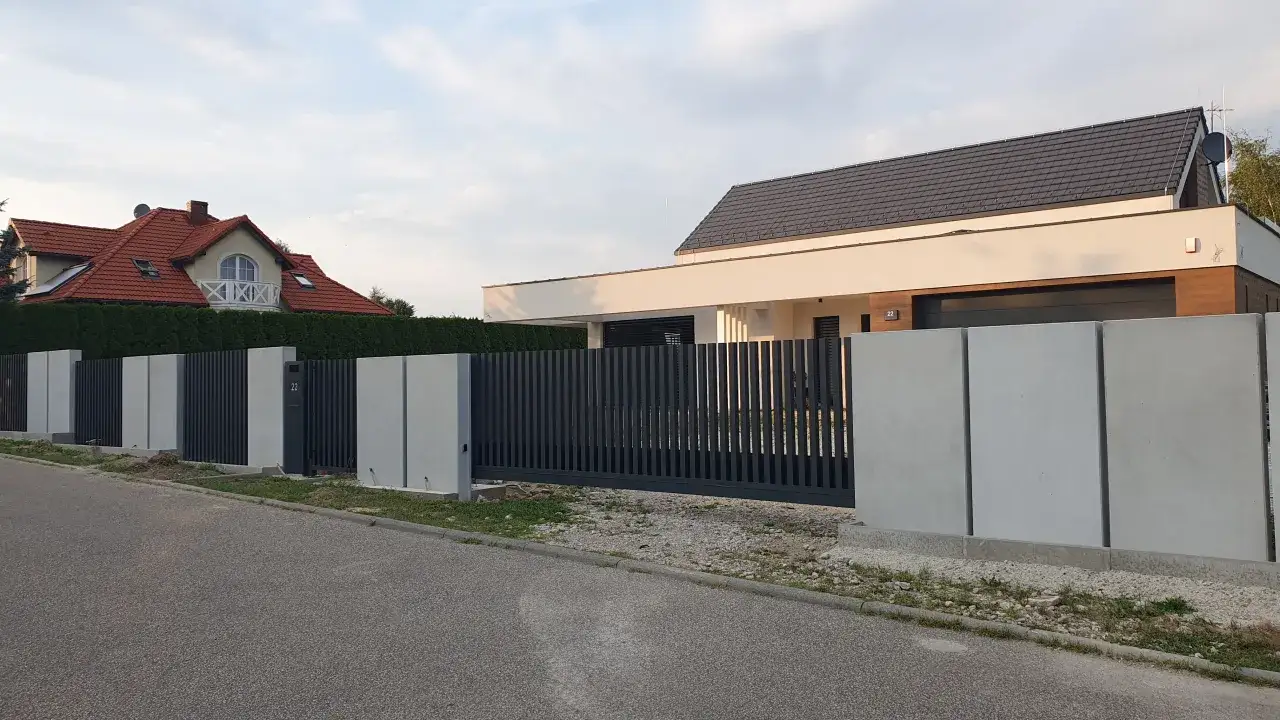 Nowoczesne ogrodzenie z beton architektoniczny i ciemnoszarymi panelami, otaczające posesję z domem.