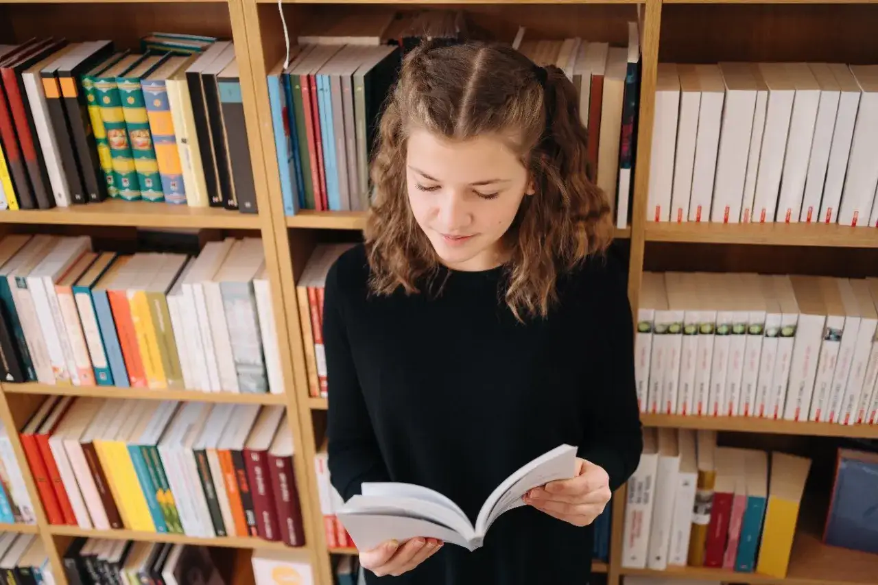Dziewczynka z warkoczykami czyta książkę na tle regałów pełnych literatury. Może to być streszczenie "Skarbczyka" lub innej lektury.