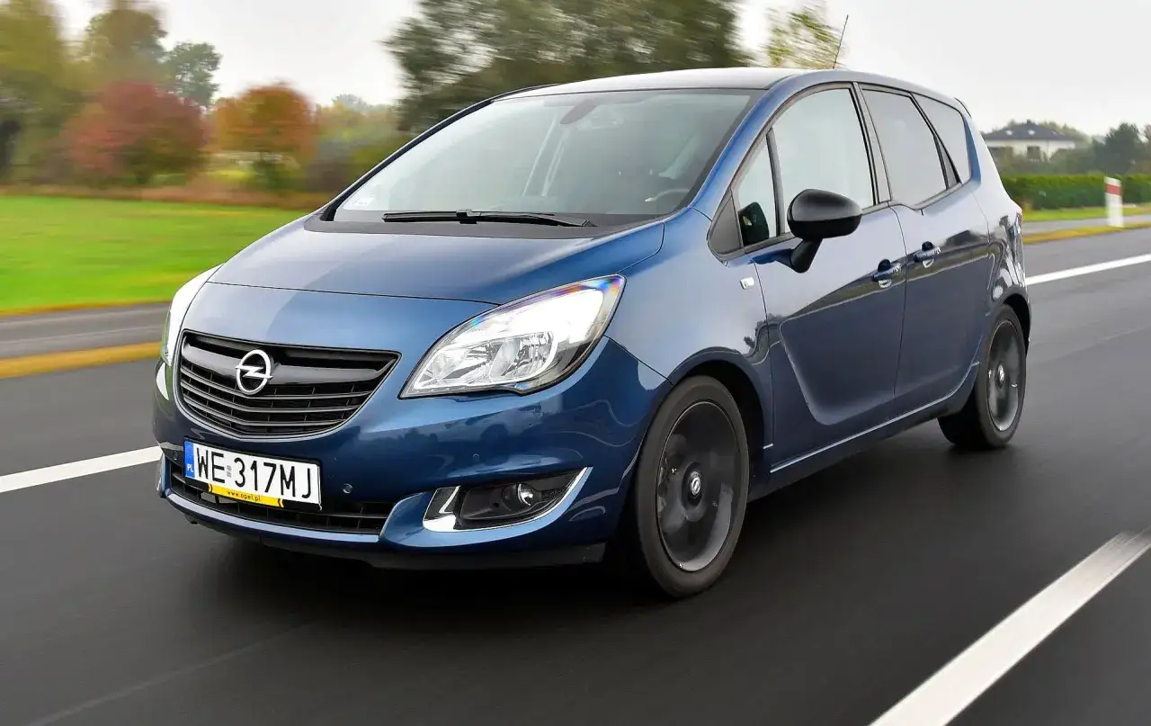 Niebieski Opel Meriva B 1.4 Turbo pędzi po drodze, otoczony jesiennym krajobrazem.