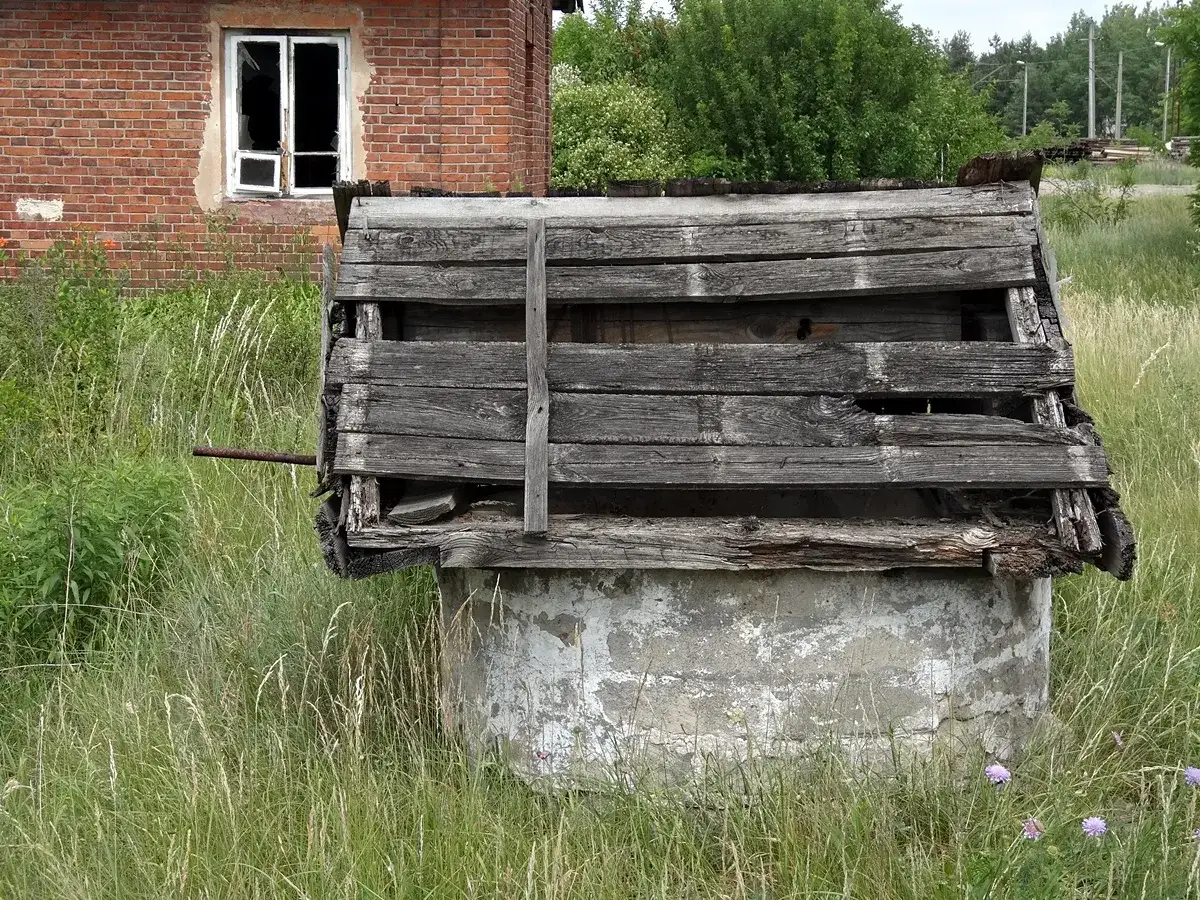 Zrujnowana, biesiadna kapela przy głębokiej studzience. Drewniany dach zniszczony, obok ceglany budynek.