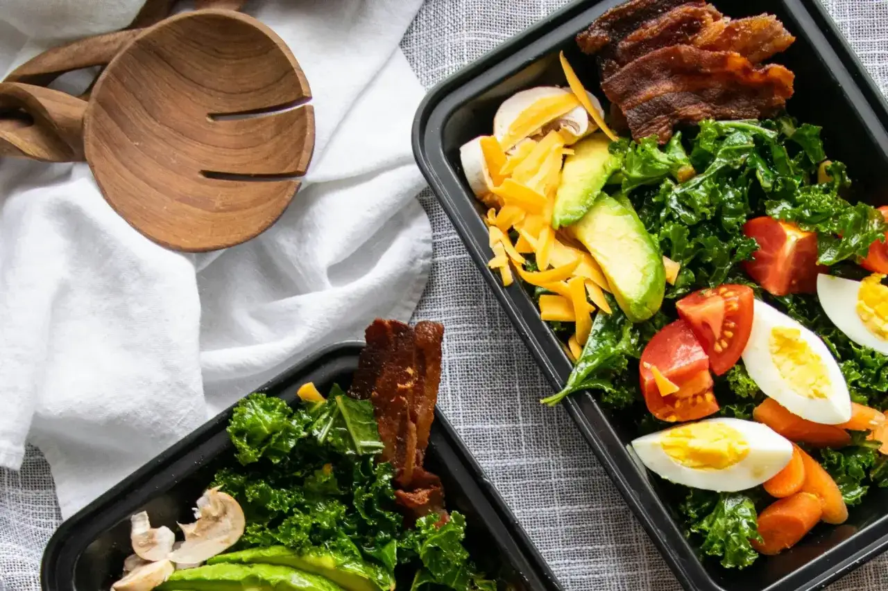 Zwei Behälter mit leckeren low carb meal prep Salaten, gefüllt mit Grünkohl, Avocado, Tomaten, Eiern, Käse und Speck.
