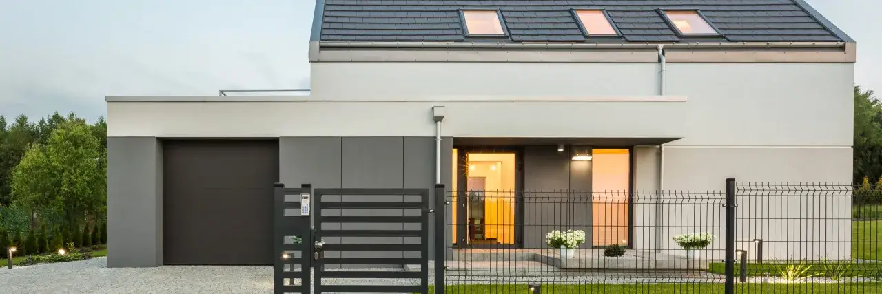 Nowoczesny dom z garażem i furtką, ale bez bramy wjazdowej. Ogrodzenie z siatki i metalowych przęseł otacza posesję.