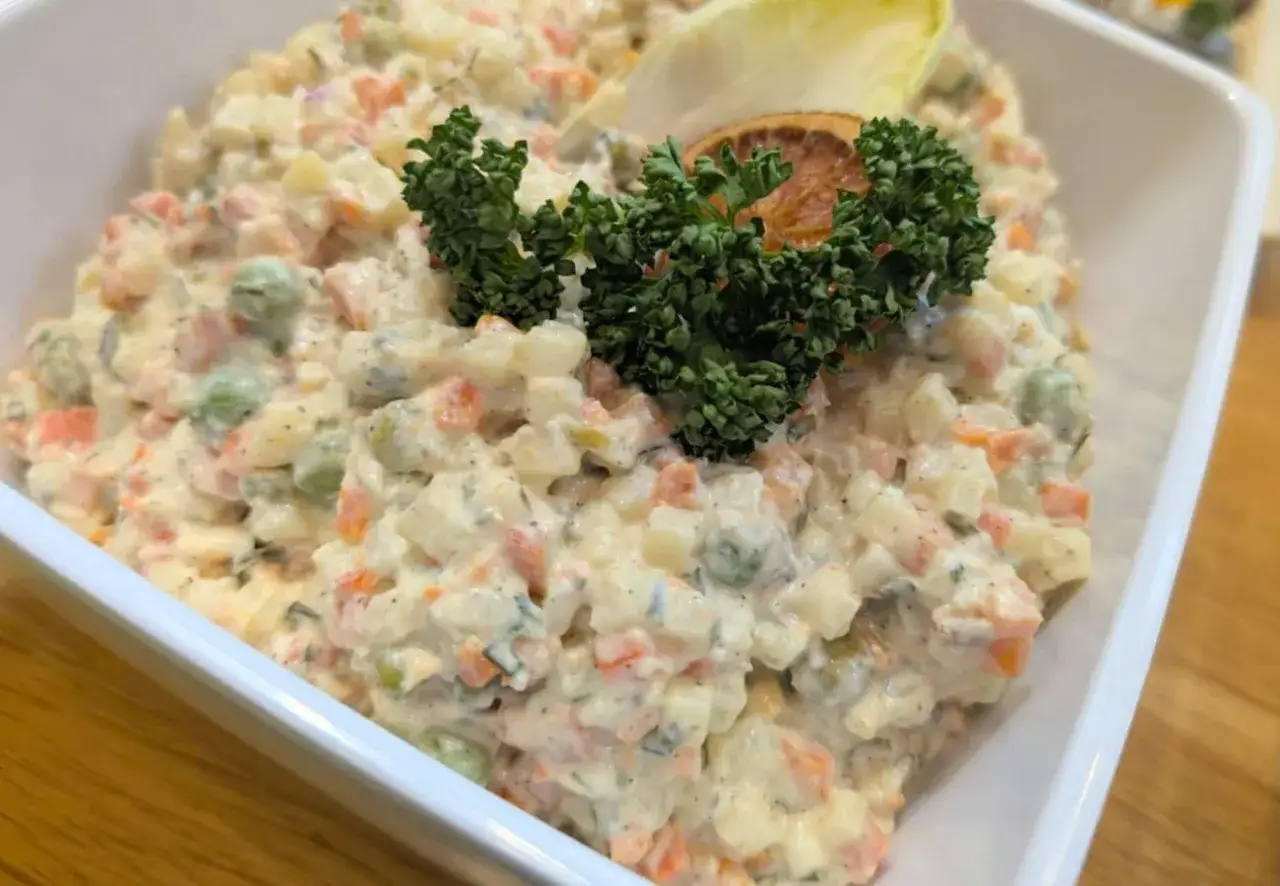 Sałatka jarzynowa w białej misce, udekorowana natką pietruszki i plasterkiem suszonej pomarańczy.