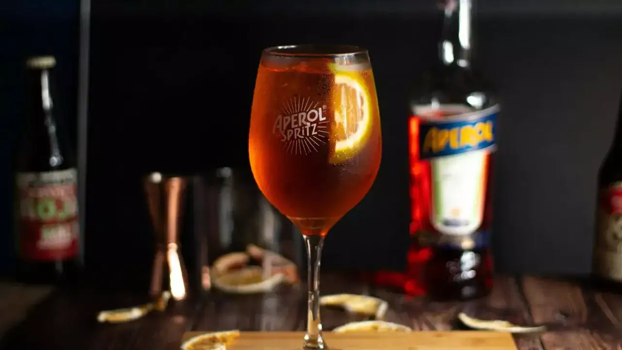 Szklanka Aperol Spritz z plasterkiem pomarańczy, butelka Aperol i akcesoria barmańskie.