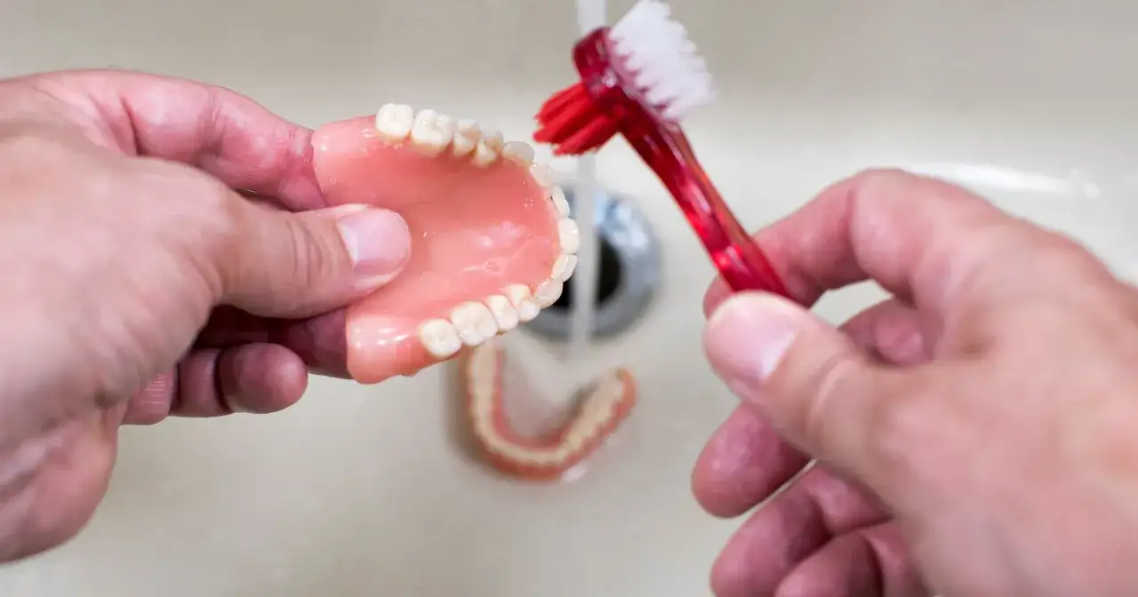 Ręce czyszczą protezę zębową pod bieżącą wodą, używając czerwonej szczoteczki. To prosty sposób, jak wyczyścić protezę zębową domowym sposobem.