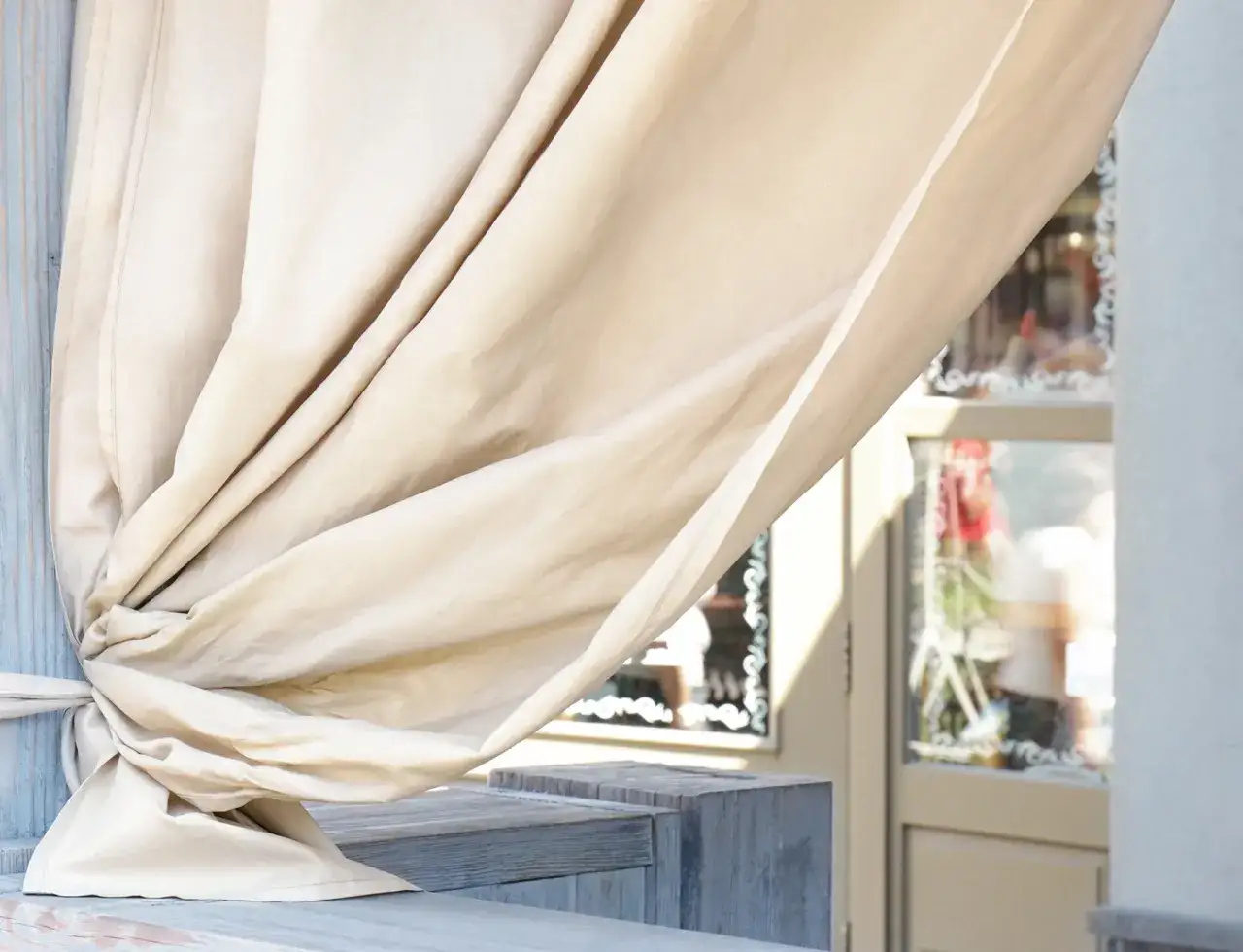 Beżowe zasłony na tarasie, zawiązane na drewnianej belce, tworzą przytulną przestrzeń. Idealne rozwiązanie na to, na czym powiesić zasłony na tarasie.