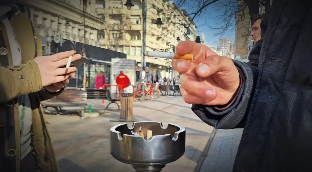 Dwie osoby palą papierosy na ulicy. W tle widać budynki i ludzi. Ceny papierosów w Albanii mogą być różne.