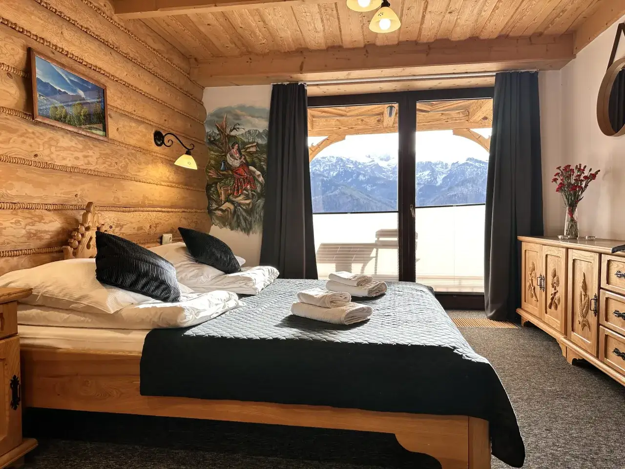 Przytulna sypialnia w stylu góralskim w Willi Kominiarski Wierch. Drewniane ściany, łóżko z czarną narzutą i widok na Tatry.