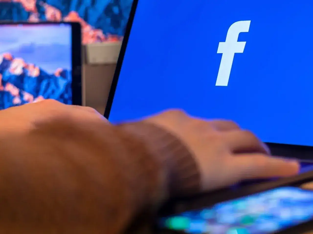 Dłoń na telefonie, obok ekranu laptopa z logo Facebooka. Dowiedz się, jak zaktualizować Facebooka na telefonie, by cieszyć się nowymi funkcjami.