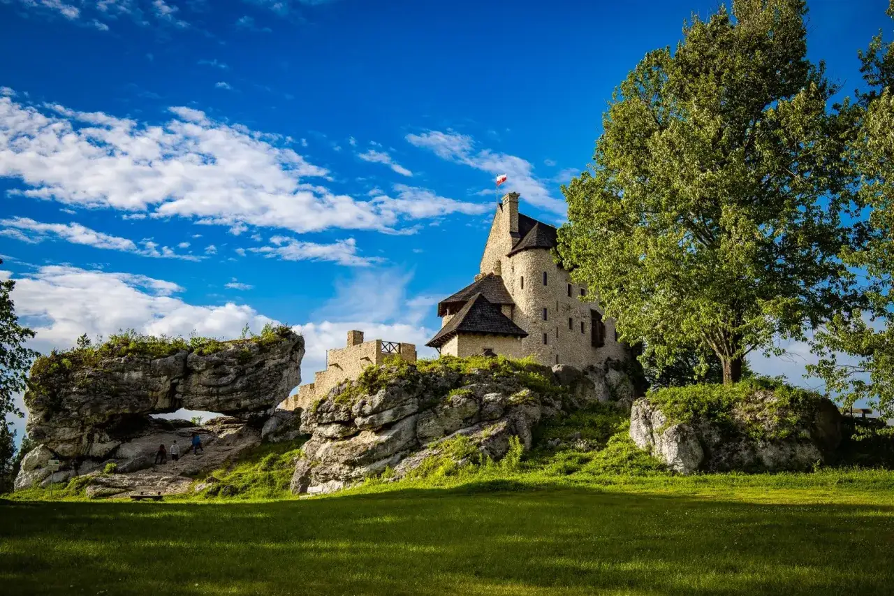 Kamienny zamek na skale, z polską flagą, w otoczeniu zieleni i skalnych formacji. W oddali widać łuk skalny, przez który przechodzą turyści. To malownicze mirów bobolice.