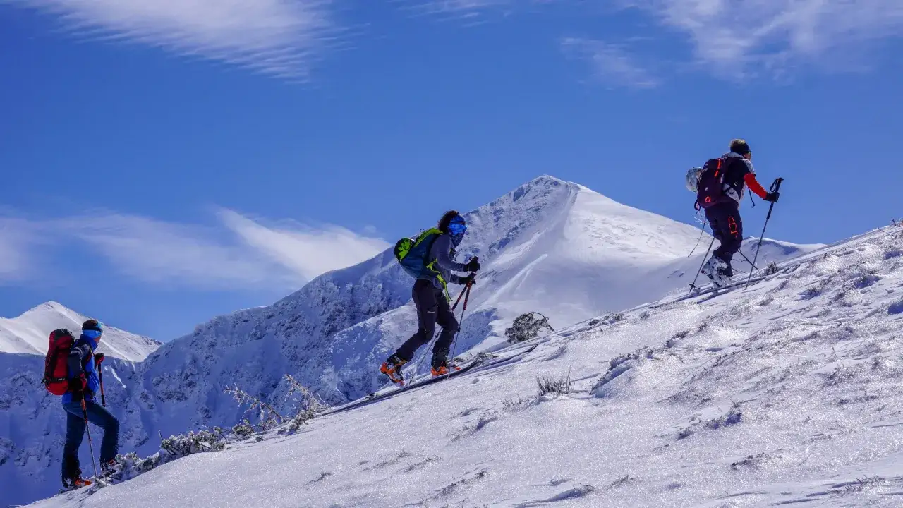 Trzy osoby na nartach skiturowych wspinają się po ośnieżonym stoku. W tle majestatyczne, pokryte śniegiem góry i błękitne niebo.