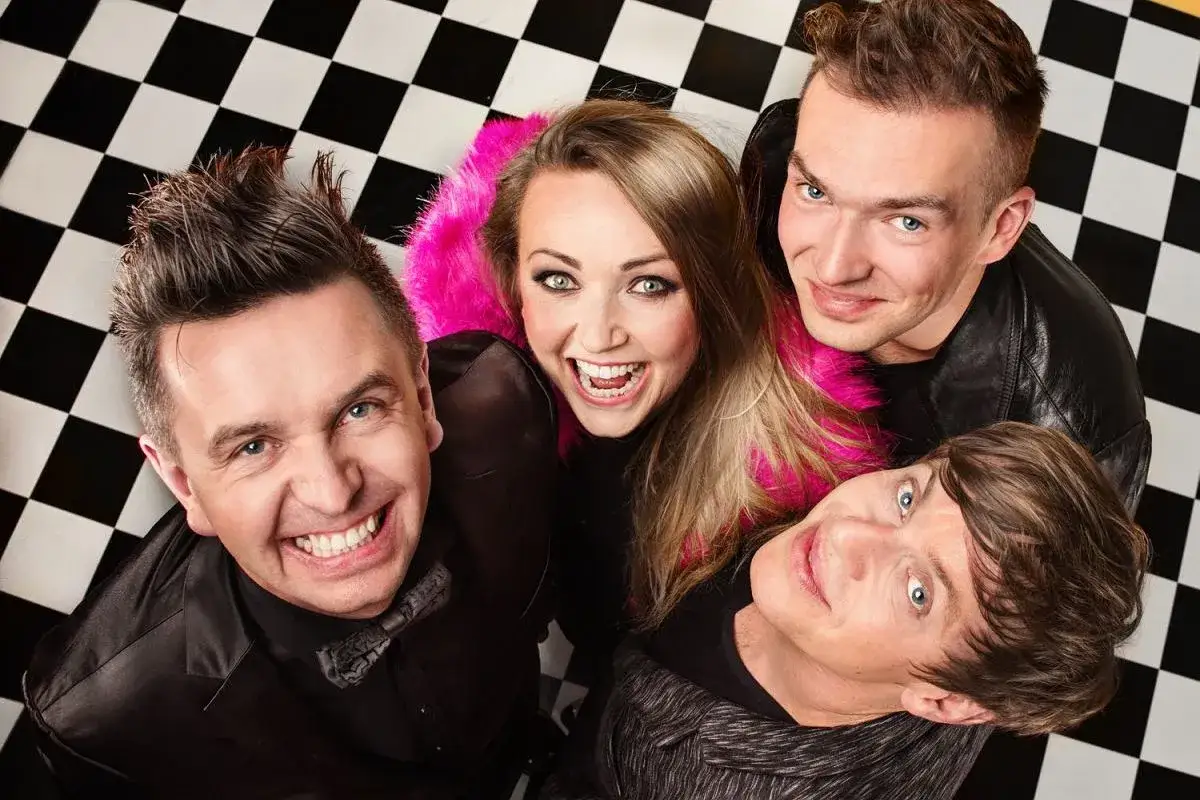Kto występuje w kabarecie Jurki? Uśmiechnięci artyści: trzech mężczyzn i jedna kobieta w różowym futerku, pozują na tle szachownicy.