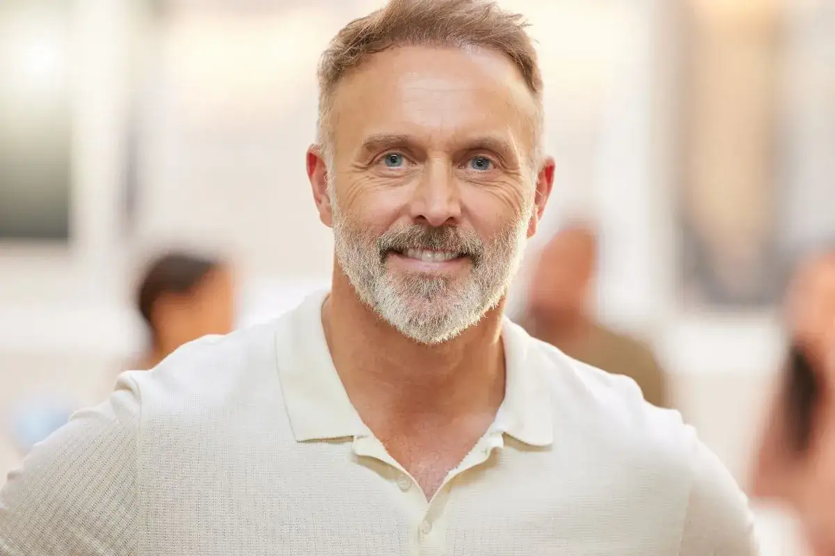 Uśmiechnięty mężczyzna w średnim wieku z siwą brodą i niebieskimi oczami, ubrany w jasną koszulkę polo.