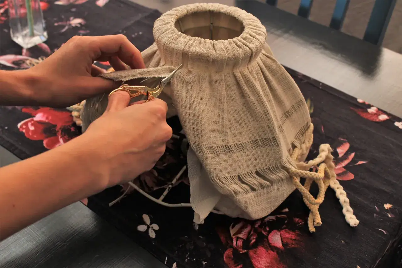 Ręce przycinają tkaninę, by zrobić abażur. Widać fragmenty materiału, nożyczki i sznurek.