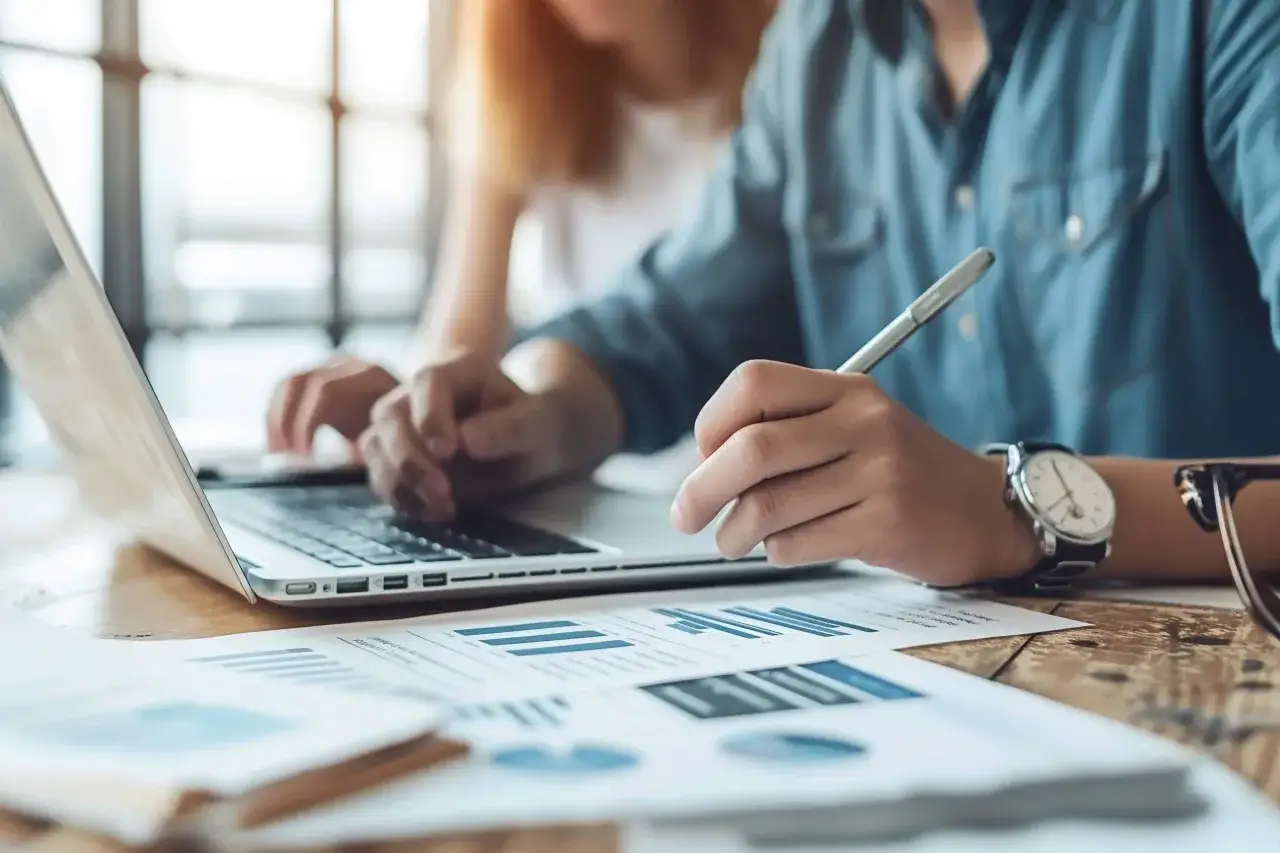Osoba w niebieskiej koszuli pisze piórem na dokumentach z wykresami obok laptopa.
