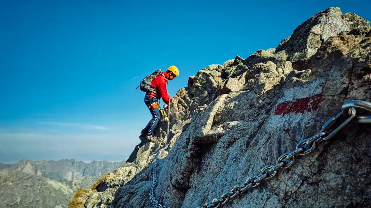 Wspinacz w kasku i czerwonej kurtce pokonuje skalną grań, używając łańcucha. Widok na tatrzańskie szczyty.