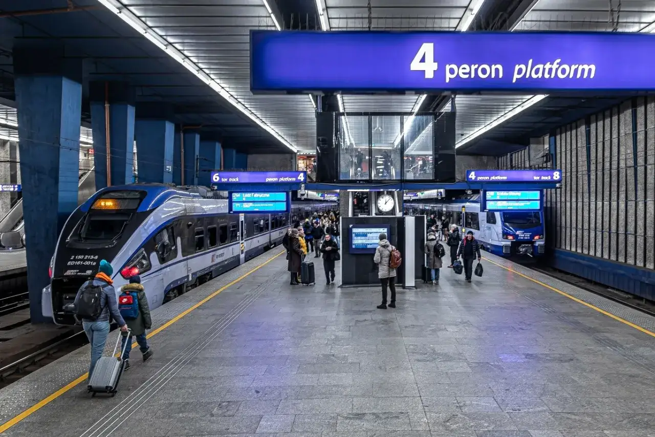 Na dworcu kolejowym ludzie czekają na pociąg. Widać nowoczesne składy i tablice informacyjne.