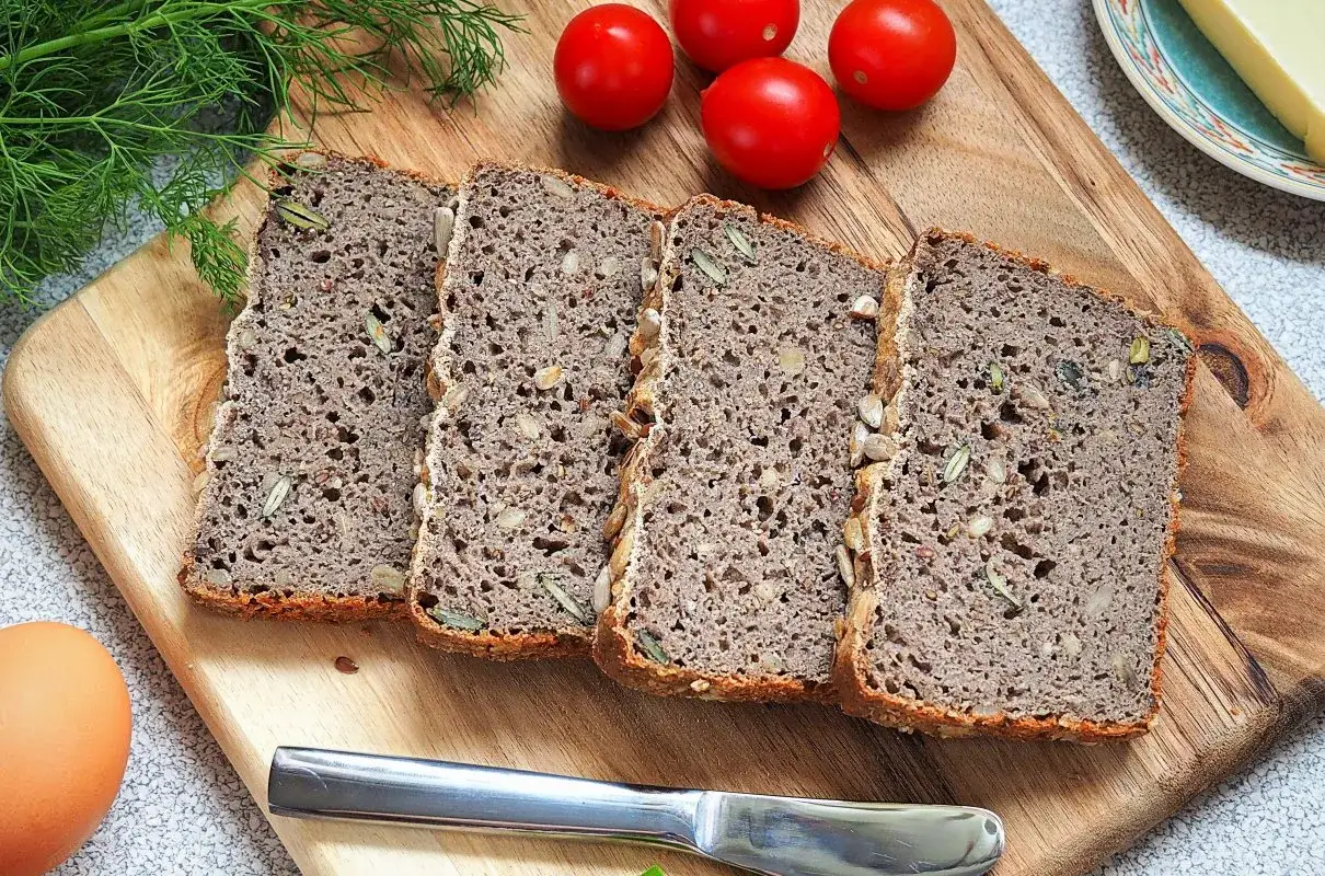Chleb gryczany bezglutenowy przepis: kromki ciemnego chleba z ziarnami na desce, obok pomidorki, jajko i nóż.