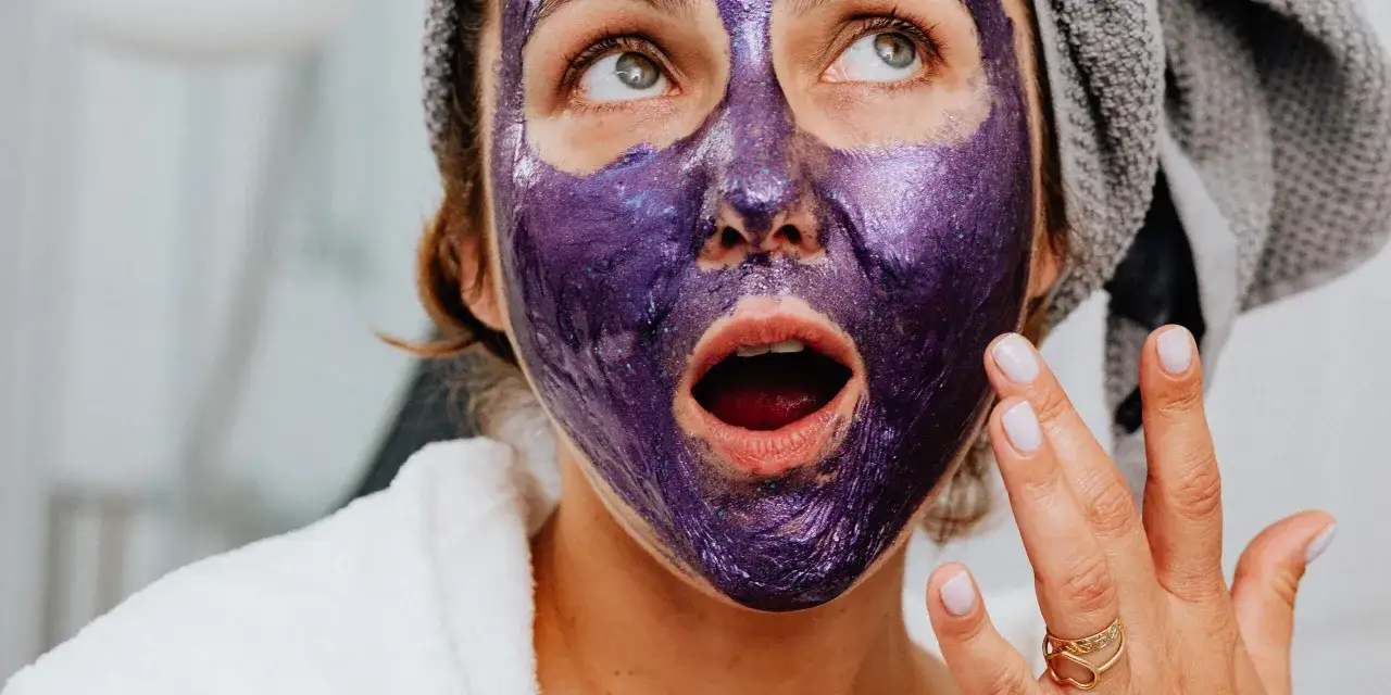 Kobieta z fioletową, błyszczącą maską na twarzy, w białym szlafroku i z ręcznikiem na głowie, cieszy się domowymi zabiegami pielęgnacyjnymi na twarz.