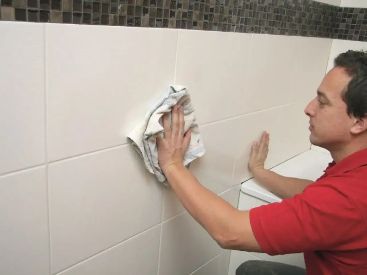 Mężczyzna czyści białe płytki na ścianie łazienki, pokazując, jak wyczyścić fugi na ścianie.