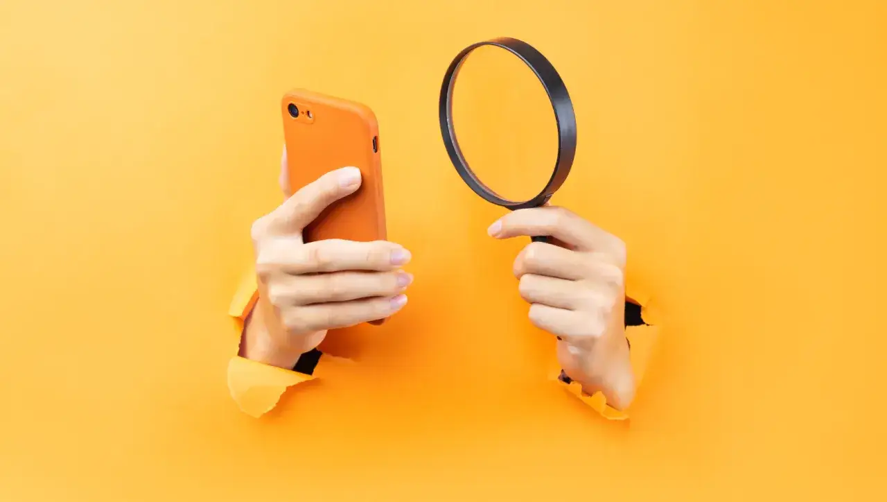 Ręce trzymające pomarańczowy iPhone i lupę, sugerujące, jak namierzyć iPhone po IMEI.