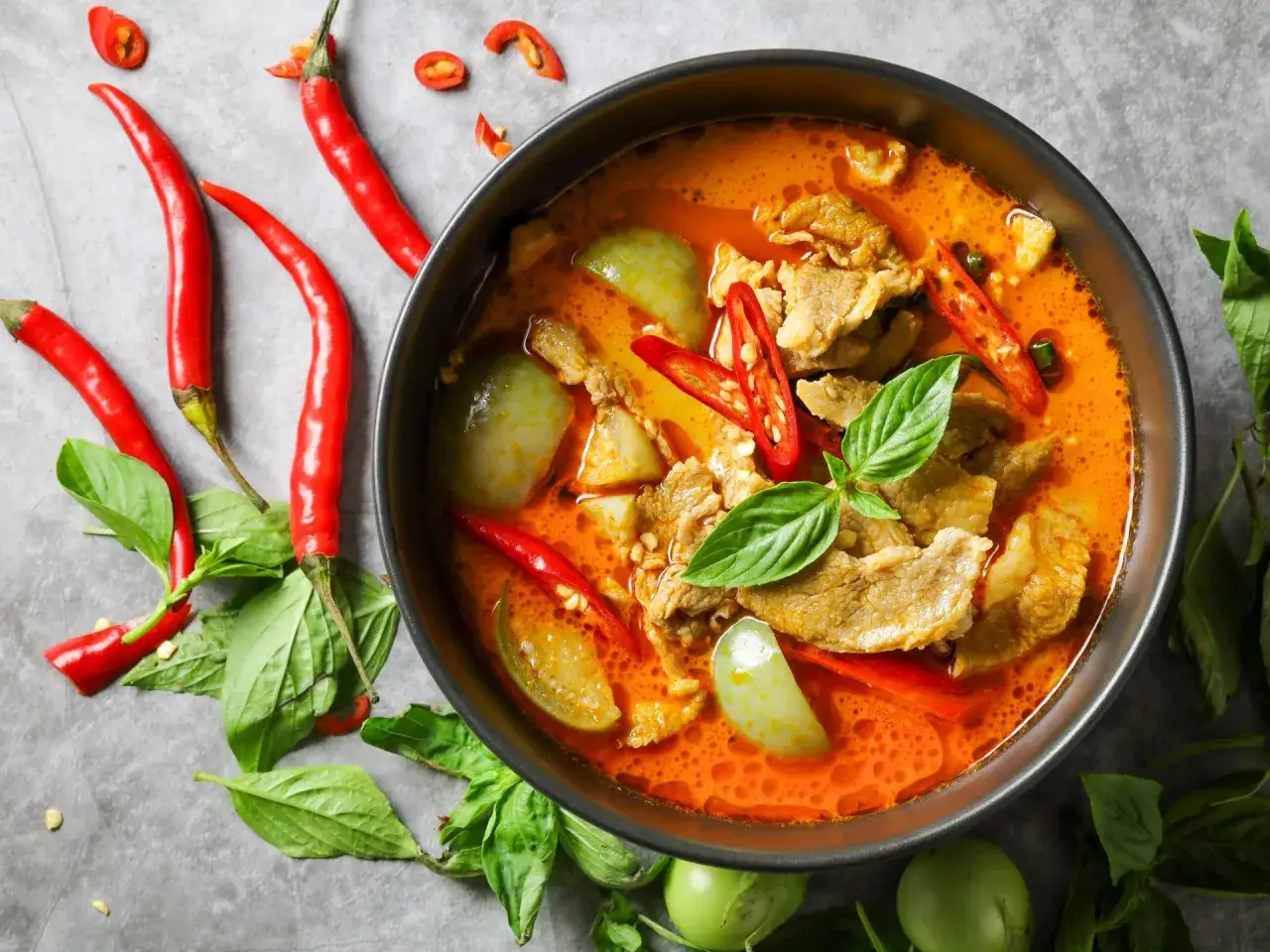 Miseczka czerwonego curry z mięsem, warzywami i papryczkami chili, otoczona świeżymi ziołami i papryczkami.