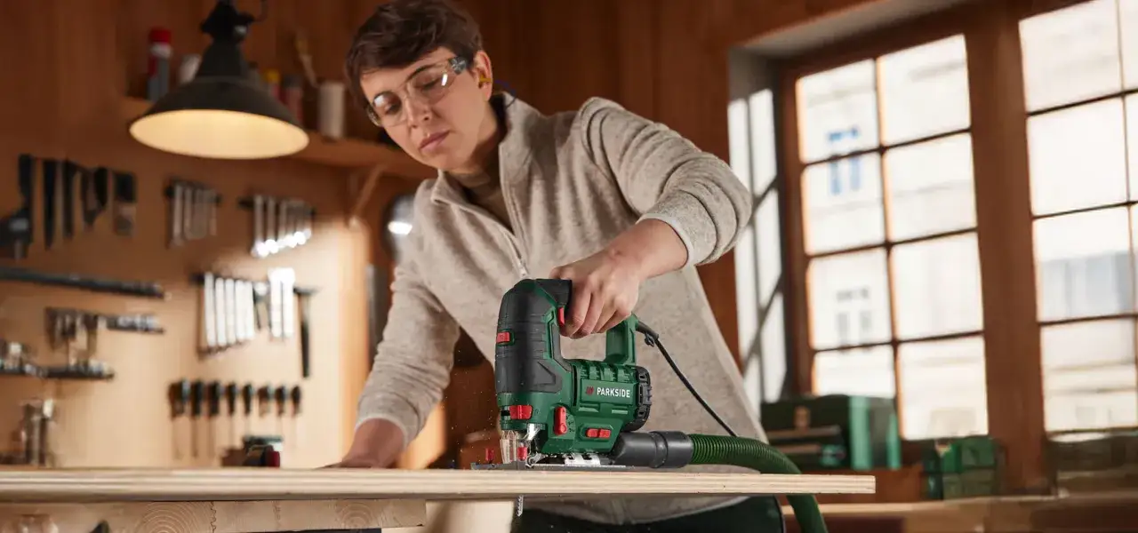 Kobieta w okularach ochronnych wykonuje cięcie drewna wzdłuż za pomocą wyrzynarki. W tle warsztat z narzędziami.